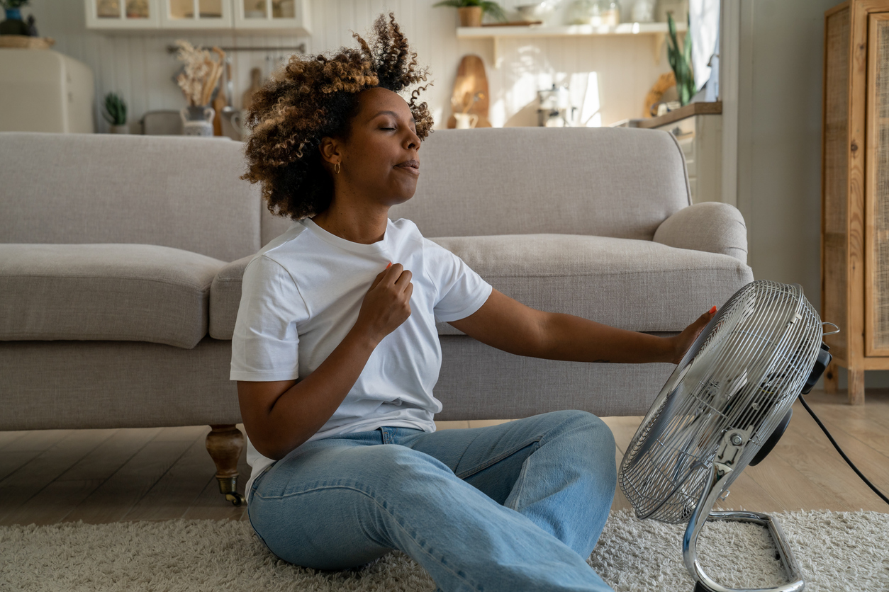Summer heat. Young woman cooling down by ventilator at home, feeling warm with high temperature during hot weather, sitting on floor in front of electric fan during heatwave